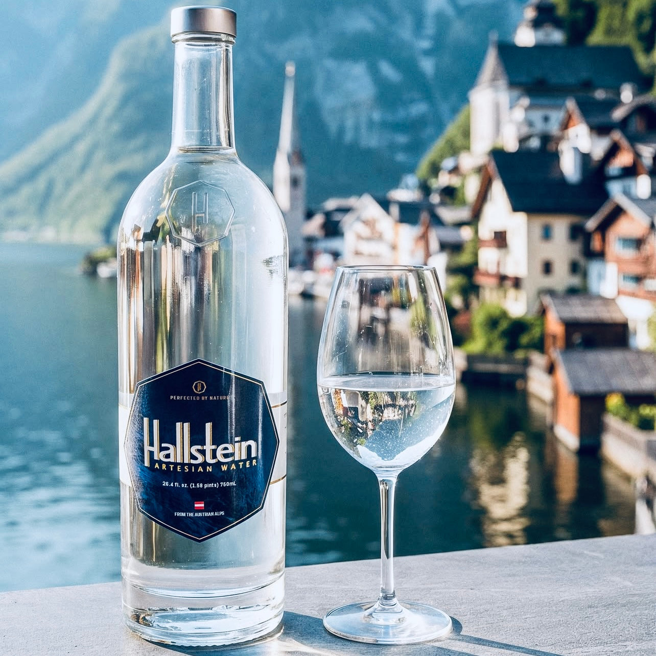 Bottle of Hallstein water with a glass against a scenic backdrop of mountains and buildings.